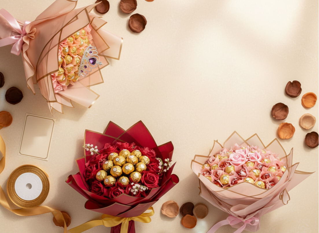 Three chocolate bouquet arrangements, two featuring pink and red roses with gold-foiled chocolates, and one with a small tiara on top, are displayed diagonally on a warm ivory surface. Scattered rose petals and small brown decorative pieces, along with a blank gift card and a spool of gold satin ribbon, serve as minimal props. The lighting is soft and warm, creating a high-end product photography style.