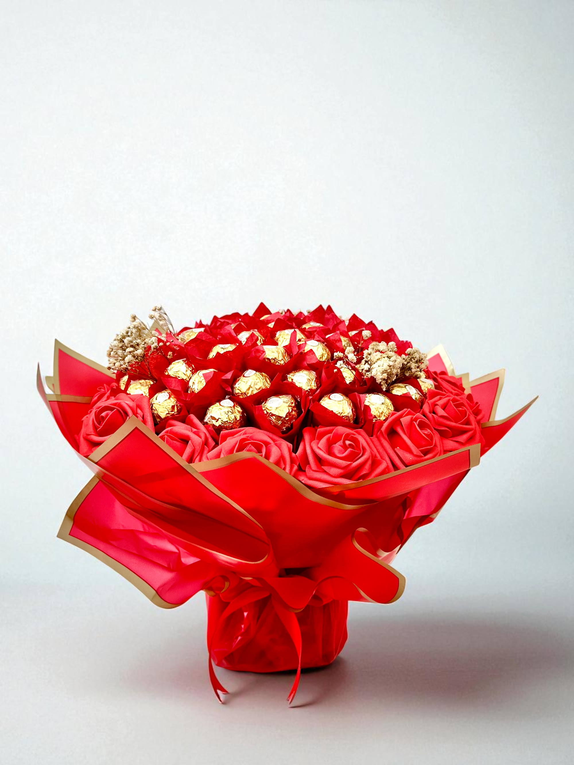 Bouquet of red roses and gold Ferrero Rocher chocolates with a smartphone displaying the same bouquet on a beige background.