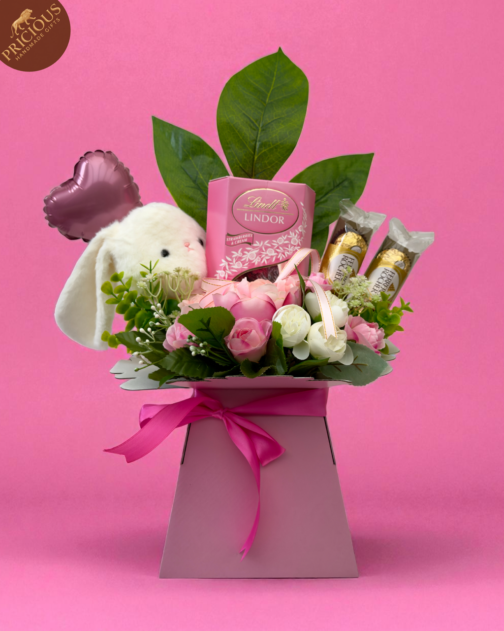 Close-up of hand-crafted gift arrangement in pink tones with foliage, chocolates and ribbon bow.